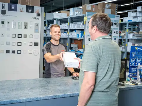 Foto von der Theke auf dem ein Mitarbeiter dem Kunden ein Produkt in die Hand drückt